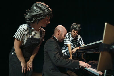 Alt-tekst: Drie performers staan rond een vleugel op een donker podium. Een man in een zwart pak speelt piano, terwijl twee anderen in gestreepte shirts en donkere tuinbroeken of broeken toekijken en over het instrument meeleunen. Ze dragen opvallende, gestileerde kapsels of pruiken. Het licht is gericht op de groep, tegen een donkere achtergrond, wat een theatraal moment suggereert.