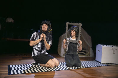 Alt-tekst: Twee performers knielen op een zwart-wit gestreept kleed op een podium. Ze dragen gestreepte shirts; één heeft een donkere doek om het hoofd geslagen en kijkt gespannen omhoog, terwijl de ander een leeg, decoratief fotolijstje voor haar gezicht houdt en opzij kijkt. Op de achtergrond staan een klein tentje van lichte stof en een witte kist, wat een speelse, theatrale setting suggereert.