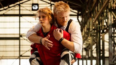 Twee personen in een industriële hal: een persoon in een rode outfit wordt van achteren vastgehouden door een man met blond haar; ze lijken samen in beweging, met metalen constructies op de achtergrond.