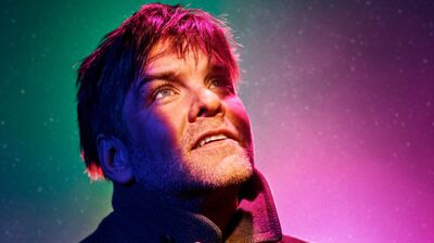 Een man met een krachtige uitstraling kijkt omhoog, verlicht door kleurrijke lichten in de achtergrond.