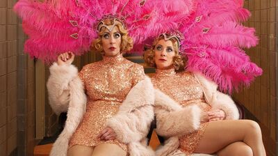 Plien & Bianca in glinsterende goudkleurige showjurken en lichtroze bontjassen zitten naast elkaar in een gang, met grote roze verenhoofden op; ze kijken opzij met een verbaasde, licht theatrale uitdrukking.