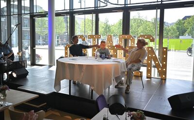 Drie mensen zitten aan een ronde tafel op een podium, omringd door grote lichtgevende letters die "STADSGASTEN" spellen.