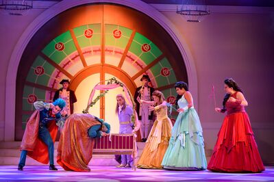 Een kleurrijke theatervoorstelling toont meerdere acteurs in uitbundige kostuums rond een wieg met roze gordijntjes. Op de achtergrond staat een grote, halfronde deur met decoratieve rozenmotieven. Twee figuren buigen zich over de wieg, terwijl anderen in lange jurken en historische kleding eromheen staan en gebaren maken. Het podium is fel verlicht met warme en paarse tinten.