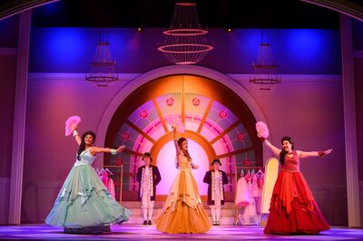 Op een kleurrijk verlicht podium staan drie vrouwen in wijde baljurken in mintgroen, goud en rood, elk met een roze pluim in de hand en in een danspose met opgeheven arm. Achter hen staat een grote halfronde deur met rozenmotieven, geflankeerd door twee personen in historische kostuums. Boven het toneel hangen meerdere kroonluchters en het decor is verlicht in roze en blauwe tinten.