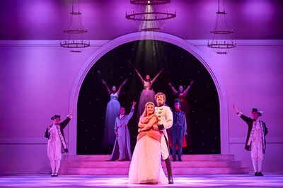 Op een sfeervol verlicht podium in paarse tinten staat een koppel centraal: een vrouw in een lichtroze baljurk en een man in een wit uniform houden elkaar vast en glimlachen. Achter hen staan meerdere performers op trappen onder een grote boog, met armen opgeheven tegen een donkere achtergrond met sterretjes. Aan weerszijden van het toneel staan twee personen in historische kostuums met uitgestrekte armen, terwijl kroonluchters boven het podium hangen.