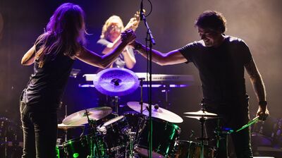 Twee muzikanten staan bij een drumstel op een podium en geven elkaar een stevige handdruk boven de drums. Beiden dragen donkere kleding en worden verlicht door paars en warm podiumlicht. Op de achtergrond is een andere bandlid zichtbaar bij een toetseninstrument. De sfeer is energiek en collegiaal, alsof ze samen een optreden afsluiten.
