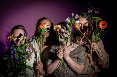 Vier personen staan dicht naast elkaar tegen een paarse achtergrond en houden boeketten met verschillende bloemen voor hun gezicht. De bloemen en bladeren werpen schaduwen over hun gezichten, waardoor hun blikken gedeeltelijk verborgen zijn.