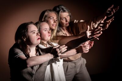 Vier personen staan schuin achter elkaar tegen een warme, bruine achtergrond en strekken hun armen vooruit in dezelfde richting. Hun gezichten zijn geconcentreerd en expressief belicht, waardoor hun blikken en handgebaren sterk naar voren komen.