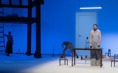 Toneelstuk met acteurs op een minimalistisch podium, met een tafel en een blauwe achtergrond.