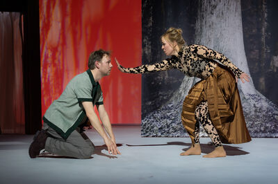 Twee acteurs op een podium: een knielende man en een staande vrouw in een dramatische pose, met een kleurrijke achtergrond.