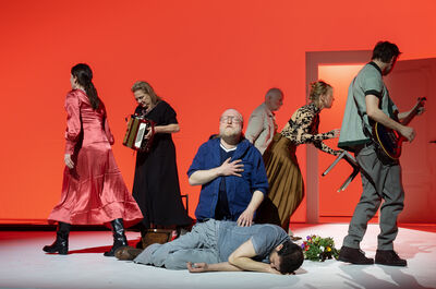 Een theateropstelling met acteurs in kostuums, waarvan één op de grond ligt terwijl anderen rondom hem staan.