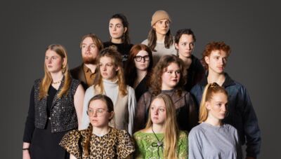 Een groep van twaalf jonge mensen staat naast elkaar voor een neutrale achtergrond, variërend in kleding en haardracht.