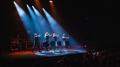 Vier percussionisten staan naast elkaar op een podium en houden hun drumstokken omhoog boven grote trommels. Felle spotlights schijnen van bovenaf op hen neer, terwijl het publiek rechts in beeld applaudisseert. Op de achtergrond staan extra percussie-instrumenten, wat zorgt voor een krachtige en theatrale concertsetting.