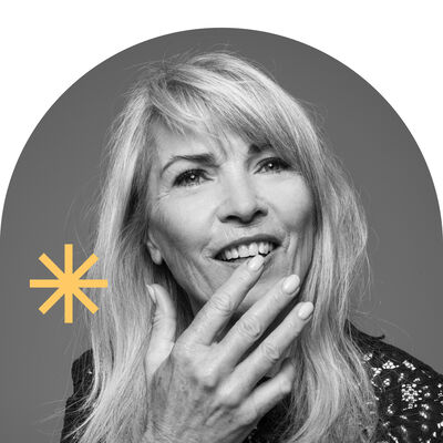 Een vrouw met lang, blond haar lacht en raakt haar gezicht aan tegen een grijze achtergrond.