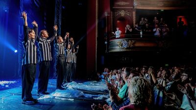 vijf performers in zwart-wit gestreepte shirts staan op een theaterpodium met hun handen omhoog en nemen applaus in ontvangst. Voor het podium klapt een volle zaal enthousiast, terwijl ook mensen op een balkon toekijken. Blauwe podiumverlichting verlicht de scène.