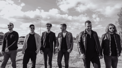 Zwart-witfoto van zes mannen die naast elkaar over een onverharde weg lopen. Ze dragen casual kleding zoals jassen, een pet en zonnebrillen, en lachen terwijl ze met elkaar praten. Op de achtergrond zijn een weg, enkele voertuigen en een lucht met grote wolken te zien.