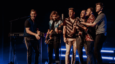 Zes mannen staan dicht bij elkaar rond een microfoon op een podium en zingen samen. Enkele van hen bespelen een gitaar en een basgitaar, terwijl op de achtergrond een toetseninstrument staat. Het podium is verlicht met blauw licht, wat zorgt voor een intieme, muzikale sfeer.