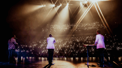 Drie muzikanten staan op een podium en kijken uit over een volle zaal. De foto is genomen van achteren, terwijl felle spotlights van bovenaf schijnen. In het publiek zijn honderden lichtjes van telefoons te zien, waardoor de zaal fonkelt als een sterrenhemel.