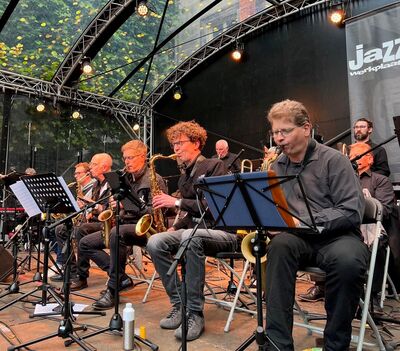 Zes muzikanten spelen op een podium tijdens een jazzconcert, met instrumenten en bladmuziek voor zich.