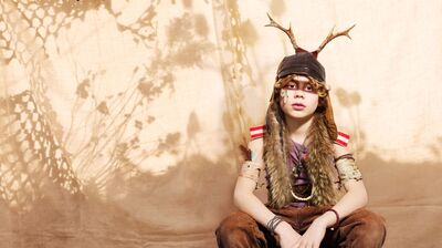 Een kind zit op de grond tegen een beige achtergrond met schaduwen van planten. Het draagt een fantasierijke outfit met een muts met geweien, een bontkraag en armbanden, en kijkt recht in de camera.