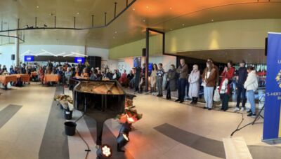 Een drukbezocht evenement in een hal met een piano, met mensen die in een rij staan en kijken naar het podium.