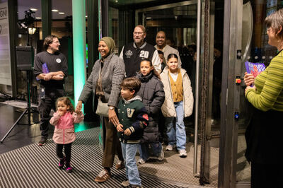 Groep mensen, waaronder kinderen en volwassenen, verlaat een gebouw en loopt naar buiten, met een feestelijke sfeer.