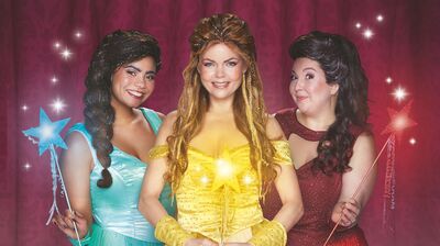 Drie vrouwen in sprookjesachtige jurken poseren glimlachend voor een roze achtergrond met sterretjes. De vrouw in het midden draagt een gele jurk met een lichtgevende ster op haar borst. De vrouw links draagt een lichtblauwe jurk en houdt een blauwe toverstaf vast, terwijl de vrouw rechts een donkerrode jurk draagt en een roze toverstaf vasthoudt.