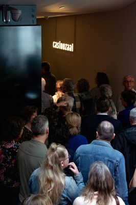 Een groep mensen in een gedimde gang, wachtend bij een lichtgevend bord met de tekst 'casinozaal'