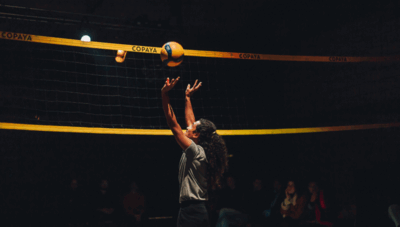 Een volleyballer met lang haar serveert de bal in een sporthal onder een net. Toeschouwers kijken toe.