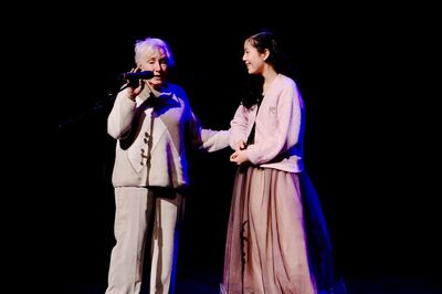 Twee vrouwelijke performers staan op een podium, met een microfoon en een lichteffect in de achtergrond.