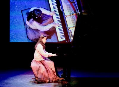 Vrouw in een lange jurk speelt piano op een podium, met een groot scherm op de achtergrond die haar afbeelding toont.