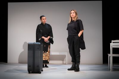 Twee vrouwen staan op een minimalistisch theaterpodium met een witte achtergrond. Links staat een vrouw in een zwarte kimono met een patroon aan de onderkant naast een grote, rechtopstaande koffer. Rechts staat een vrouw in een zwarte outfit met stevige veterlaarzen, haar handen in haar zakken. Op het podium staan verder een witte bank en een tafel, wat de scène een sobere en ingetogen uitstraling geeft.