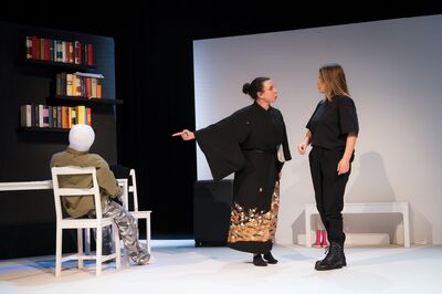 Theatrale scène op een minimalistisch podium met een witte achterwand en een zwarte boekenkast vol boeken. Links zit een opblaaspop aan een witte tafel, met de rug naar het publiek. In het midden wijst een vrouw in een zwarte kimono met patroon nadrukkelijk naar de pop, terwijl een andere vrouw in zwarte kleding en veterlaarzen naast haar staat. Een koffer en een witte bank maken deel uit van het sobere decor.