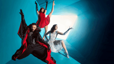 Drie dansers in theatrale kostuums poseren in een felblauwe ruimte met een helder verlichte opening: links een persoon in een zwart-rood gewaad met opgeheven arm, bovenin een danser in een rode jurk met een hoog gestrekt been, en rechts een danser in een lichte jurk die zittend een arm naar het licht uitstrekt.