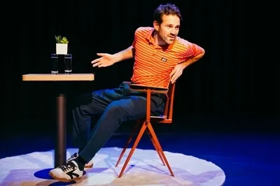 Martijn Crins zit op een oranje designstoel op een verlicht rond vloerkleed op een verder donker podium. Hij draagt een oranje-wit gestreept poloshirt en een donkerblauwe broek, en kijkt met een vragende uitdrukking opzij terwijl hij zijn armen spreidt. Naast hem staat een klein tafeltje met daarop twee glazen water en een klein wit plantenbakje met vetplantjes.