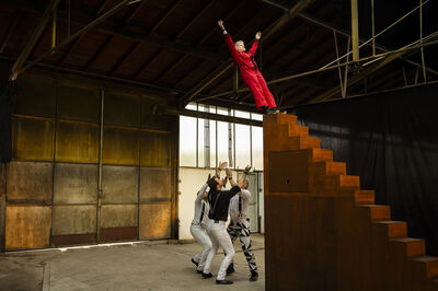 In een grote industriële hal staat een persoon in een rode overall bovenaan een hoge, trapvormige constructie met de armen omhoog, terwijl drie personen beneden hun handen uitstrekken alsof ze haar proberen op te vangen.