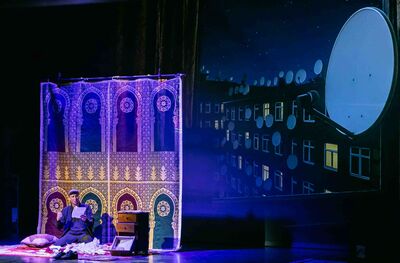 Theatrale scène waarin Jawad Es Soufi op een kleed op de grond zit tussen papieren en een klein houten kastje, voor een decor met oosterse patronen; rechts is een projectie te zien van een nachtelijk flatgebouw met meerdere satellietschotel op het dak.