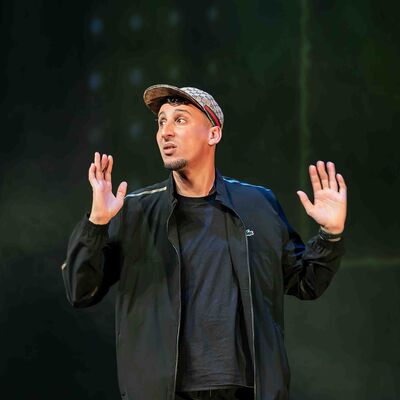 Jawad Es Soufi met pet en zwarte jas staat op een podium met Donkere achtergrond, kijkt opzij en houdt beide handen omhoog alsof hij iets benadrukt tijdens een optreden.