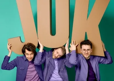 Drie jonge mannen in paarse outfits staan naast elkaar tegen een heldergroene achtergrond. Ze houden boven hun hoofden drie grote letters van bruin karton vast die samen het woord 'JUK' vormen. De linker man draagt een paars overhemd, de middelste een paars suède jasje en de rechter man een paars bomberjack en een ronde bril.