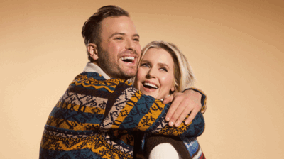 Een lachende man en vrouw omhelzen elkaar innig tegen een warme beige achtergrond. De man draagt een kleurrijke gebreide trui en slaat zijn armen om de vrouw heen, terwijl zij glimlachend tegen hem aan leunt. De sfeer is warm en vrolijk.