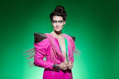 Vrouw in een opvallend roze kostuum met schouder- en fransemotieven, posed voor een groene achtergrond.