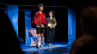 Twee performers staan op een verlicht podium tijdens een theatervoorstelling. De persoon links draagt een rode cape over een gestreept shirt en zwarte shorts, terwijl de ander een gestreept shirt en donkere broek draagt en een platte afbeelding van een beer vasthoudt. Op de achtergrond staan decorstukken in blauwe tinten die een bos suggereren en een open kist. Op de voorgrond zijn vaag de silhouetten van toeschouwers te zien.