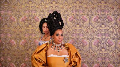 Twee vrouwen poseren voor een achtergrond met goudkleurig, bloemrijk behang. Op de voorgrond staat een vrouw in een goudkleurige jurk met opvallende sieraden en een hoge, gevlochten haarcreatie; ze kijkt recht in de camera met een zelfverzekerde blik. Achter haar staat een andere vrouw met krullend haar en een tiara, gekleed in een elegante jurk, die met gesloten ogen naar beneden kijkt. De styling en kleding doen denken aan een historische of koninklijke setting.