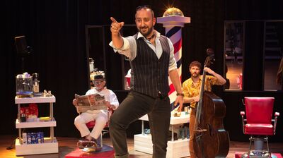 Een performer in een vest en overhemd staat op het podium, omgeven door muzikanten en vintage barberstoelen.