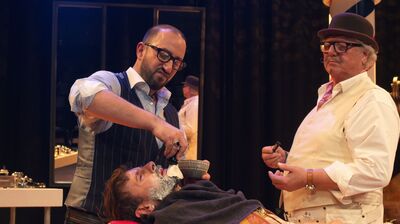 Een barber geeft een scheerbeurt aan een klant terwijl een tweede man toekijkt.