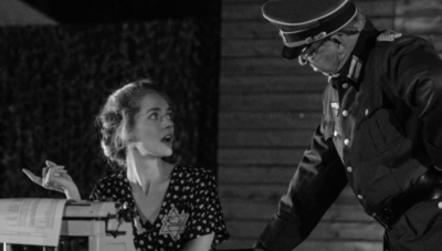 Een vrouw in een stippenjurk kijkt verbaasd naar een man in een uniform terwijl ze bij een bureau zit.