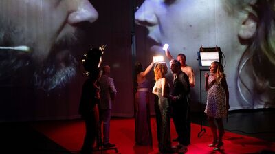 Toneelopvoering met acteurs die een intense interactie hebben onder projecties van gezichten op de achtergrond.