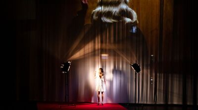 Een vrouw in een witte jurk staat onder een schijnwerper op een podium, met schaduwen die op de achtergrond zichtbaar zijn.