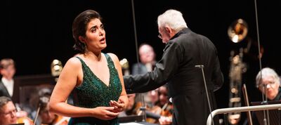 Zangeres in een groene jurk zingt terwijl een dirigent de orkestleden aanstuurt op een podium.