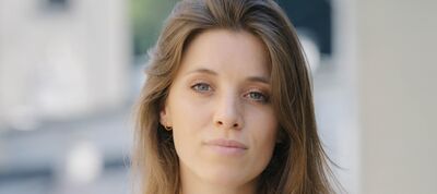 Portret van een vrouw met lange, bruin haar en neutrale gezichtsuitdrukking, staande in een stedelijke omgeving.